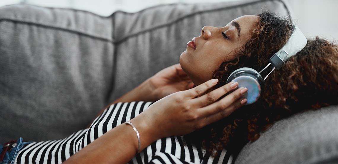 Someone laying on a grey couch wearing white over-ear, noise-cancelling headphones.