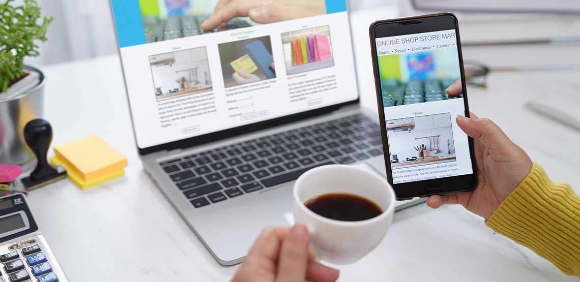 A person holding a phone and a cup of coffee with an open laptop in the background showing a business website.