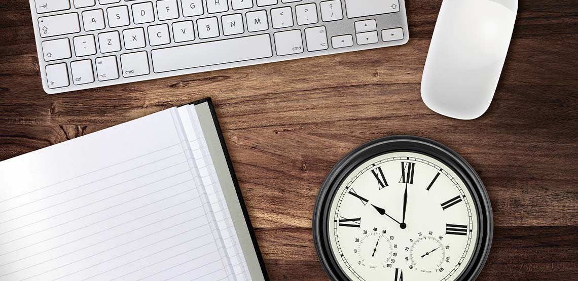 A flat lay of an open notebook,. a clock, a keyboard and a mouse against a wooden table.